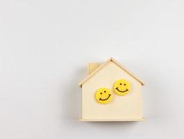 eben legen von hölzern Modell- Haus mit zwei Gelb Kreis lächelnd Gesichter auf Weiß Hintergrund mit Kopieren Raum. glücklich heim, glücklich Familie Konzept. foto
