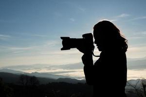 Schattenbild des jungen Fotografen, der eine Kamera mit Berglandschaft hält