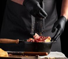 Koch im schwarz Uniform und Latex Handschuhe Würze roh Hähnchen Fleisch im ein schwarz Besetzung Eisen Bratpfanne, Kochen foto