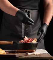 Koch im schwarz Uniform und Latex Handschuhe Würze roh Hähnchen Fleisch im ein schwarz Gusseisen braten schwenken foto