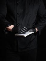 Koch im schwarz Uniform und Latex Handschuhe halten ein leer Notizbuch und ein schwarz hölzern Bleistift foto