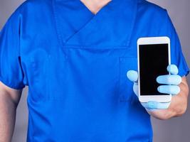 Arzt im Blau Uniform und Latex Handschuhe zeigen ein Weiß Smartphone mit ein leer schwarz Bildschirm foto