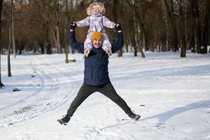 Vater halt Kind auf Schultern während Springen zusammen beim sonnig eisig Winter Tag im das Park. Papa und Tochter Liebe. foto