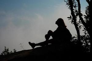 Silhouette eines Wanderers, der auf Berggipfel mit blauem Himmelhintergrund sitzt foto