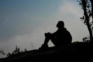 Silhouette eines Wanderers, der auf Berggipfel mit blauem Himmelhintergrund sitzt foto