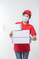 Bild eines bewussten jungen Zustellers mit roter Kappe, leerem T-Shirt, einheitlichen Gesichtsmaskenhandschuhen, die mit leerem weißen Karton isoliert auf hellgrauem Hintergrund Studio stehen foto