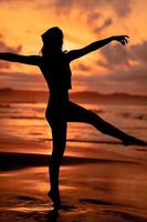 ein Ballerina mit ein Silhouette gestalten führt aus Ballett Bewegungen sehr flexibel auf das Strand mit das Wellen abstürzen foto