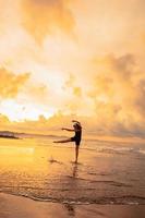 ein asiatisch Frau im ein schwarz Kleid führt aus Ballett Bewegungen schön auf das Strand mit Wellen und Wolken hinter ihr foto