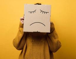 ein Frau im ein braun Sweatshirt mit ein Box auf ihr Kopf steht auf ein Gelb Hintergrund. traurig Grimasse, Stress und Depression Konzept. mental Gesundheit foto