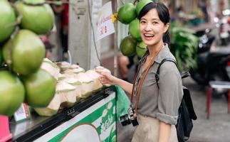 glücklich jung asiatisch Frau Rucksack Reisender Stand im Vorderseite von Kokosnuss Saft Geschäft beim China Stadt, Dorf Straße Essen Markt im Bangkok, Thailand. Reisender Überprüfung aus Seite Straßen. foto