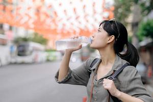 glücklich jung asiatisch Frau Rucksack Reisender Trinken ein kalt Wasser beim China Stadt, Dorf Straße Essen Markt im Bangkok, Thailand. Reisender Überprüfung aus Seite Straßen. foto