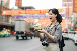 glücklich jung asiatisch Frau Rucksack Reisender Trinken ein Kokosnuss Saft beim China Stadt, Dorf Straße Essen Markt im Bangkok, Thailand. Reisender Überprüfung aus Seite Straßen. foto