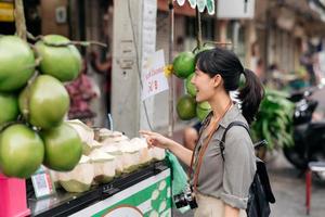 glücklich jung asiatisch Frau Rucksack Reisender Stand im Vorderseite von Kokosnuss Saft Geschäft beim China Stadt, Dorf Straße Essen Markt im Bangkok, Thailand. Reisender Überprüfung aus Seite Straßen. foto