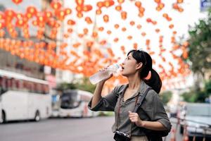 glücklich jung asiatisch Frau Rucksack Reisender Trinken ein kalt Wasser beim China Stadt, Dorf Straße Essen Markt im Bangkok, Thailand. Reisender Überprüfung aus Seite Straßen. foto