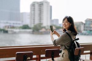 jung asiatisch Frau Rucksack Reisender mit Handy, Mobiltelefon Telefon im ausdrücken Boot Seebrücke auf Chao Phraya Fluss im Bangkok. foto