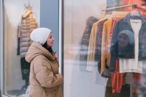 ein stilvoll jung Frau im ein Hut und Mantel im das Stadt sieht aus in das Fenster von Kleidung Shops foto