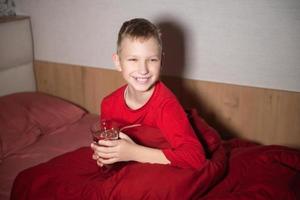 ein glücklich Junge ist Sitzung im ein rot Bett unter ein Decke mit ein Glas von Wasser foto