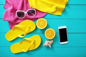 Sommer- Zubehör auf Blau hölzern Hintergrund. Gelb Flip Flops, Handtücher, Sonnenbrille, Handy, Mobiltelefon Telefon und Orangen. spotten oben und oben Aussicht foto