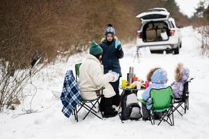 jung Frau mit Kinder im Winter Wald auf ein Picknick. Mutter und drei Kinder gegen Sie Auto mit öffnen Stamm. foto