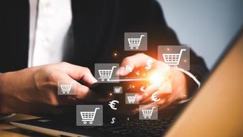 Geschäftsmann halten Telefon und Laptop benutzt zum online Einkaufen durch Websites auf das Internet, selektiv Fokus von klein Einkaufen Taschen in der Nähe von Spielzeug Einkaufen Wagen mit Laptop auf Hintergrund. foto
