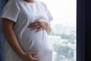 Foto von glücklich asiatisch schwanger Frau Sitzung im Bett und berühren ihr Bauch beim Zuhause . mit klein Schuhe zum das ungeboren Baby im das Bauch