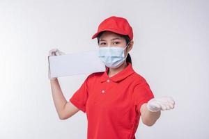 Bild eines bewussten jungen Zustellers mit roter Kappe, leerem T-Shirt, einheitlichen Gesichtsmaskenhandschuhen, die mit leerem weißen Karton isoliert auf hellgrauem Hintergrund Studio stehen foto