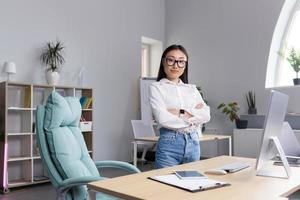 asiatisch Frau Geschäftsfrau im das Büro beim das Schreibtisch. gekreuzt ihr Waffen, sieht aus beim das Kamera, lächelt foto