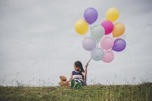 kleines Mädchen mit einem Teddybär und Luftballons auf Wiesenfeld foto