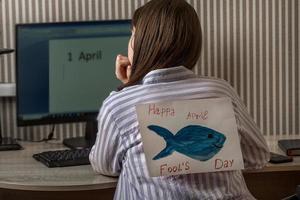 eine junge frau mit einem papierfisch von hinten drinnen arbeitet an einem computer, auf dem die aufschrift erster april, aprilscherz, witze und witze im büro übereinander stehen foto