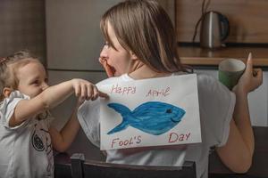 ein fröhliches kleines mädchen klebt ihrer mutter zu hause in der küche einen papierfisch auf den rücken, eine junge frau frühstückt, in der hand hält sie eine tasse tee. lustige familienwitze, aprilscherzfeier foto