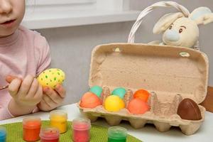 Mädchen ist Konzentration auf Zeichnung Muster auf ein Ostern Ei, Nächster zu es ist ein Container mit gemalt Eier und ein Korb mit ein Ostern Hase foto