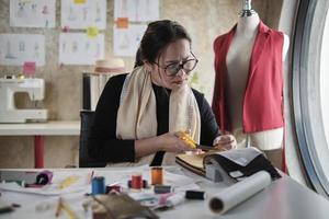 asiatische modedesignerin mittleren alters arbeitet im studio, schneidet und wählt stoff- und fadenfarben mit zeichnungsskizzen für kleiderdesign-kollektionen. professioneller Boutique-Schneider-Unternehmer. foto