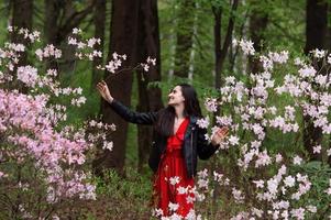 ein jung Mädchen im ein schwarz Jacke und ein rot Kleid steht in der Nähe von ein Weiß Rhododendron foto