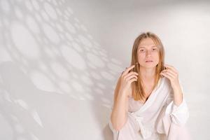 nachdenklich Süss Frau posieren auf Licht Hintergrund mit Licht Kunst Schatten im ein durchscheinend Weiß Shirt, traurig Stimmung foto