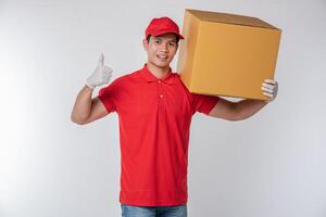 Bild eines glücklichen jungen Zustellers in roter Kappe, leerem T-Shirt, Uniform, stehend mit leerem braunem Karton, isoliert auf hellgrauem Hintergrund Studio foto