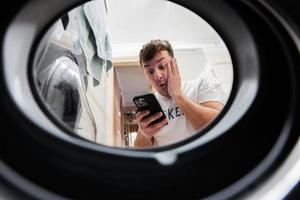 Mann mit Handy, Mobiltelefon Telefon, Aussicht von Waschen Maschine innen. männlich tut Wäsche Täglich Routine. foto