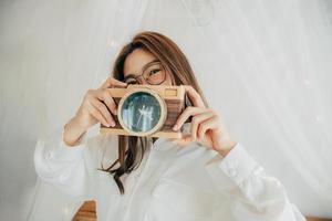 Porträt einer glücklichen asiatischen Schönheit, die im weißen Hemd lächelt und auf dem Bett posiert. Fotografieren von einer Uhr, die wie eine Fotokamera aussieht. Menschen Lifestyle-Konzept. Platz kopieren. Blick in die Kamera. foto