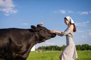 ein jung Mädchen im ein Kuh Weide steht im ein Kleid und ein Kopftuch und Schlaganfälle ein Kuh foto