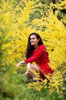 ein Mädchen im ein rot Kleid sitzt in der Nähe von ein Busch mit Gelb Blumen und sieht aus in das Entfernung foto