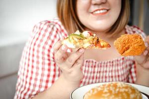 ein asiatisch Fett Frau ist Essen Pizza und gebraten Hähnchen auf das Sofa im ihr Haus. das Konzept von verbrauchen Essen Das Ursachen Krankheit ist nicht gut zum Gesundheit foto