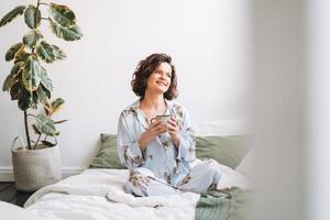 jung lächelnd Frau mit Brünette lockig Haar im Blau Pyjama mit Tasse von Tee im Hände Sitzung auf Bett beim Zuhause foto