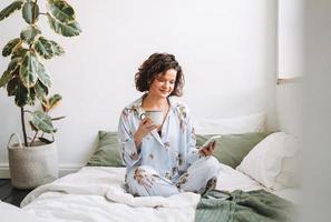 jung Brünette Frau mit lockig Haar im Pyjama mit Handy, Mobiltelefon auf Bett beim Zuhause foto