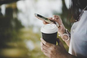 junge Frau, die Wegwerfkaffeetasse hält, während Textnachrichten durch Smartphone im Freien