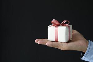 Hand hält eine kleine Geschenkbox mit rosa Band auf schwarzem Hintergrund