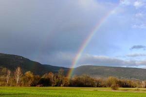 Regenbogen auf dunkel Regen Wolken Über Grün Felder mit Bäume und Berge foto