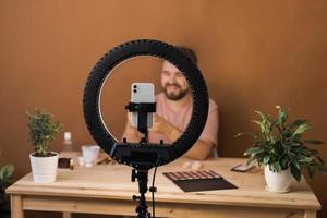 männlich Schönheit Blogger mit bilden Produkt Aufzeichnung Video beim heim. mit Ring Lampe und Smartphone. vlog kosmetisch Produkte und Vielfalt Konzept foto