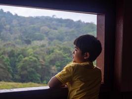 Junge, der aus dem Fenster blickt und den grünen Wald betrachtet foto