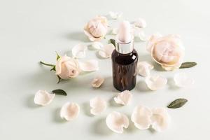 kosmetisch natürlich Öl oder Serum zum aktiv Selbstpflege im ein Flasche mit ein Tropfer auf ein Weiß Hintergrund unter das Knospen und Blütenblätter von ein Weiß Rose. foto