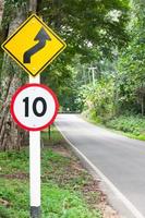 Selektive Geschwindigkeitsbegrenzung Verkehrszeichen 10 und kurvenreiches Straßenwarnsymbol für sicheres Fahren auf der Landstraße im Wald mit Bergblick, zurückhaltend foto