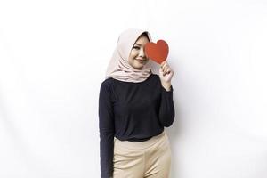 ein glücklich jung asiatisch Muslim Frau tragen ein Hijab fühlt sich romantisch Formen Herz Geste ausdrücken zärtlich Gefühle und halten ein rot herzförmig Papier foto